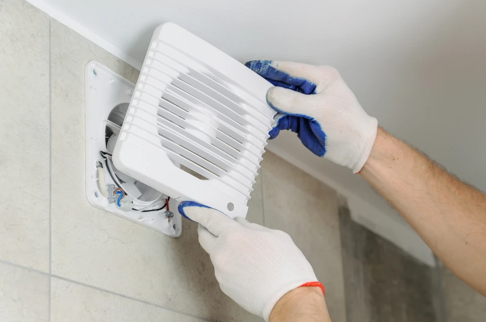 Technicien électricien professionnel en tenue de travail bleu installant une unité centrale de VMC (ventilation mécanique contrôlée) dans les combles ou un grenier d'une maison, avec accès visible aux gaines de ventilation. L'image montre clairement l'installation d'un système complet de renouvellement d'air pour le logement.